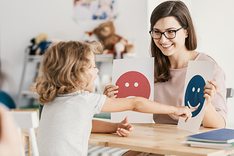 This is a stock photo from Shutterstock. An adult woman is sitting at a table facing a young girl. The woman is holding up two pictures. One picture is a "happy" face, and the other picture is a "sad" face. The young girl is pointing at one of the pictures.