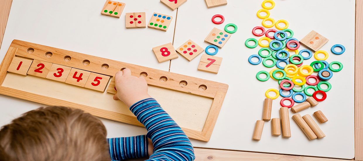 This is a stock photo from Shutterstock. The image is overlooking a white table with colorful puzzle pieces spread across the table. The puzzle pieces include number, shapes, and dominos. A child's hands are arranging the puzzle pieces on a puzzle board.